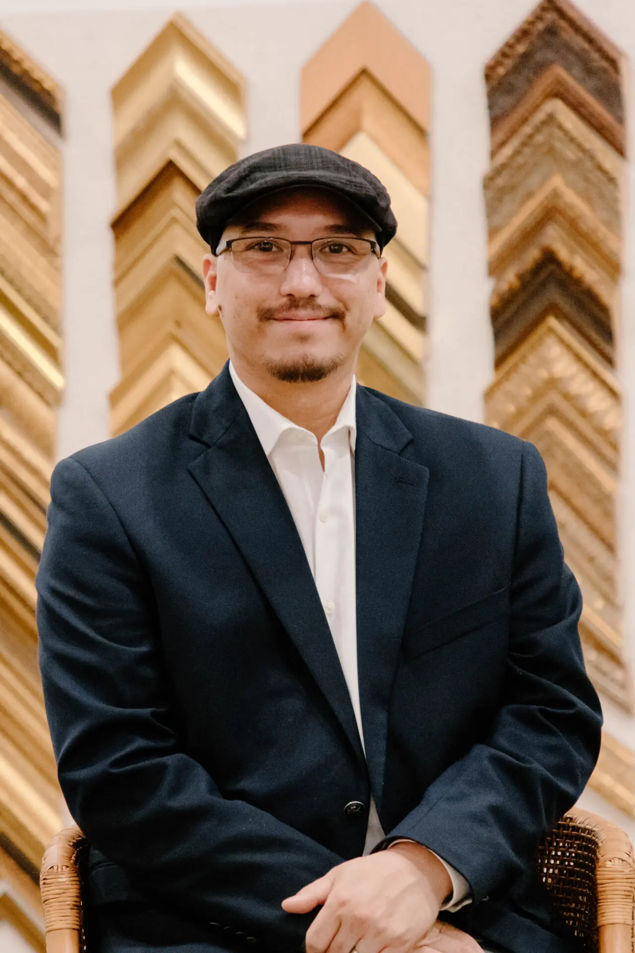 Man with glasses and a black hat sitting with his hands crossed smiling at camera